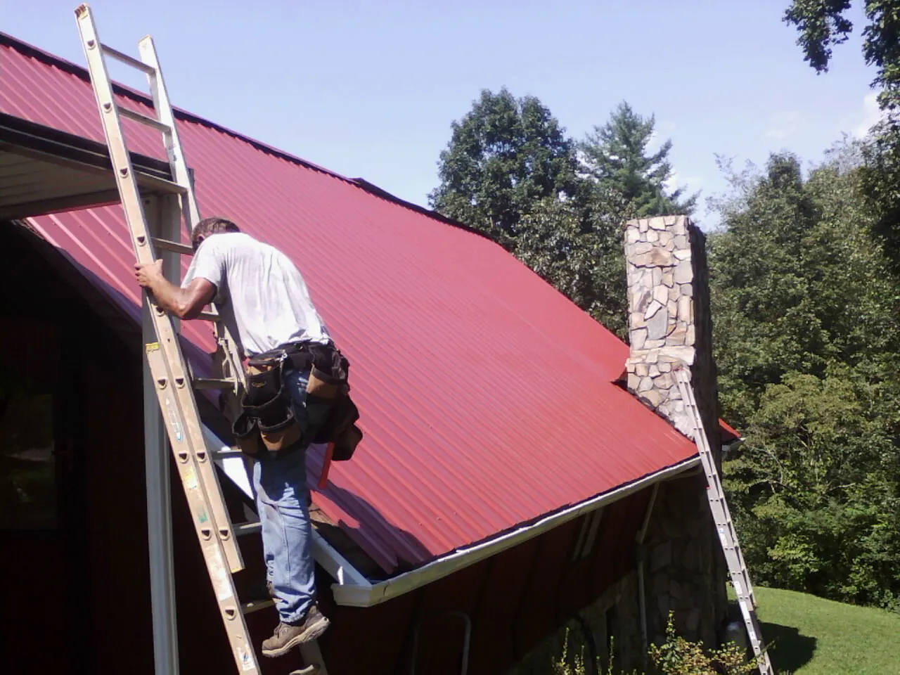 Roofer on a ladder beside a red metal roof and stone chimney — Soffit & Fascia Repair by Roofing Brothers in Southington